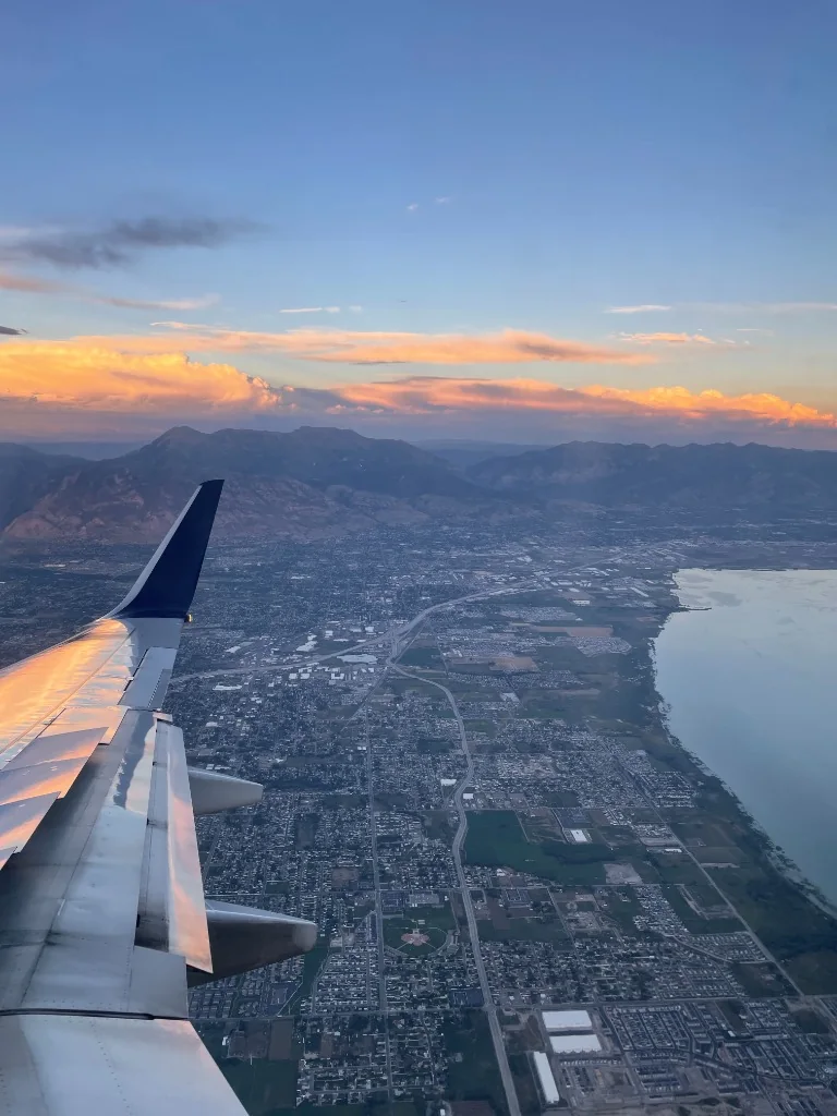 Airplane wing above a city, mountains, and clouds lit by sunset